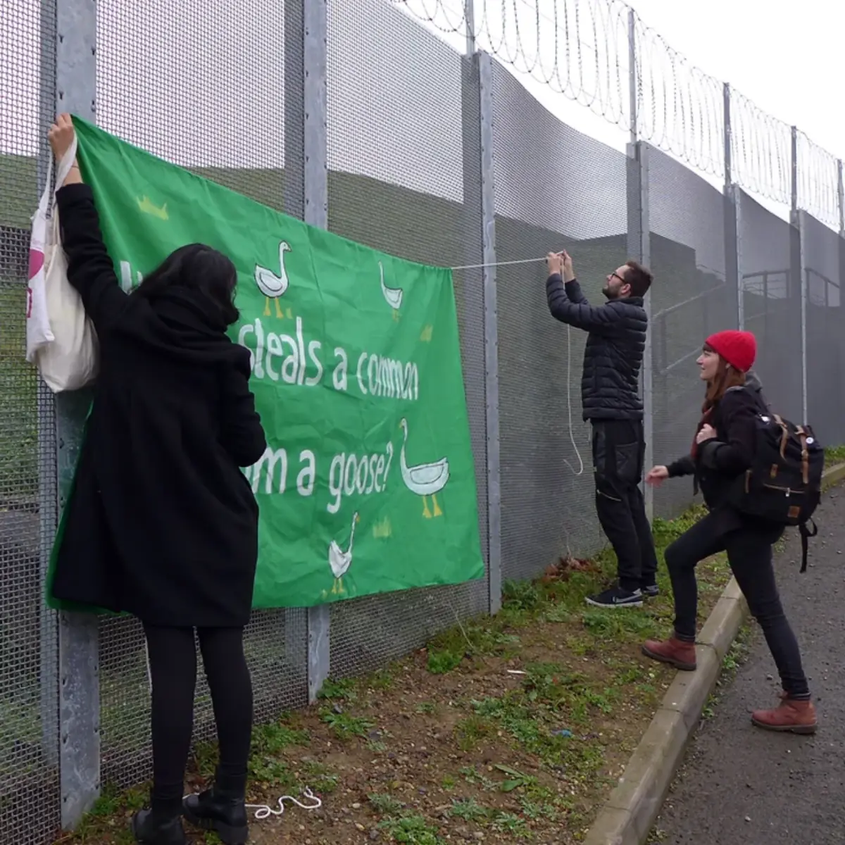 Three campaigners tie a green banner to the reservoir's steel fence, reaching up above their heads.