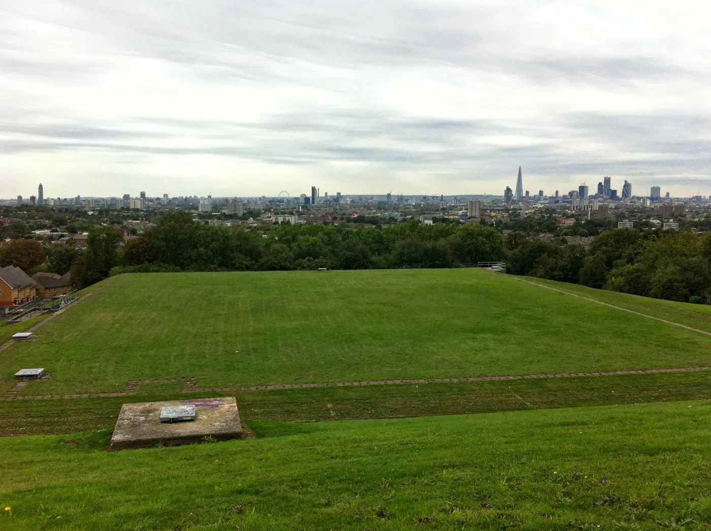 An aerial view of the reservoir: a large flat green rectangle ringed by fencing, surrounded by terraced streets, with the City of London on the horizon.