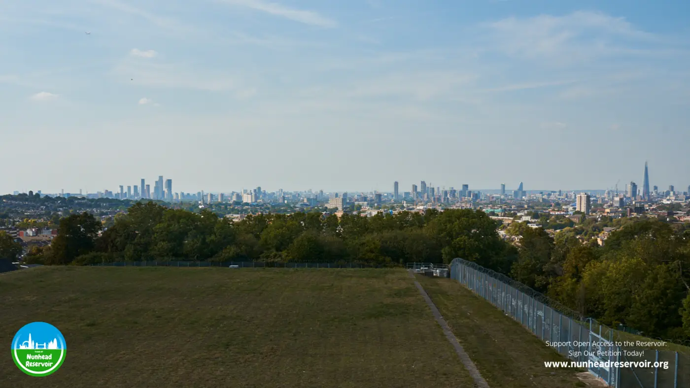 The flat grass top of the reservoir in daylight, its fence running the length of the far edge, with the skyline beyond.